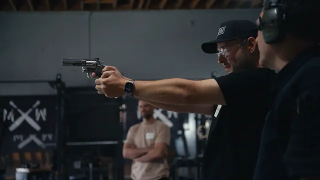 Josh aims a prop revolver during Dutch Merrick's firearms safety course, with Tommy watching behind him.