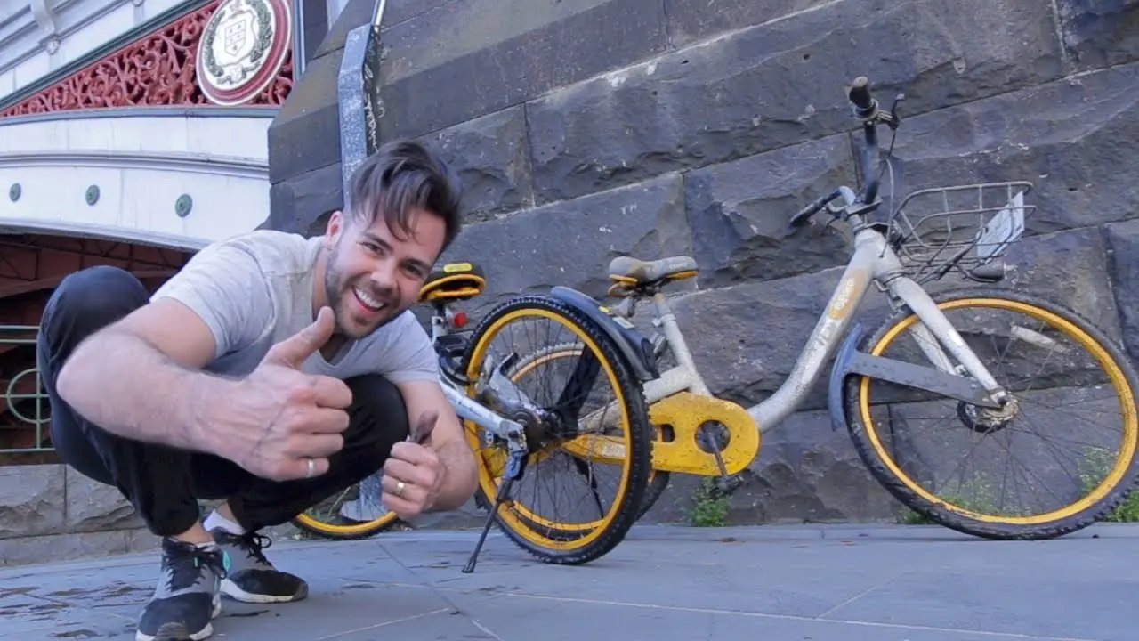 Thumbnail for the video The O Bike Rabbit Hole: Tommy Jackett crouching and giving a thumbs up beside a yellow oBike.